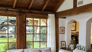 Amplia sala de estar con vigas de madera, ventilador de techo y ventanas grandes que ofrecen vistas al exterior.
