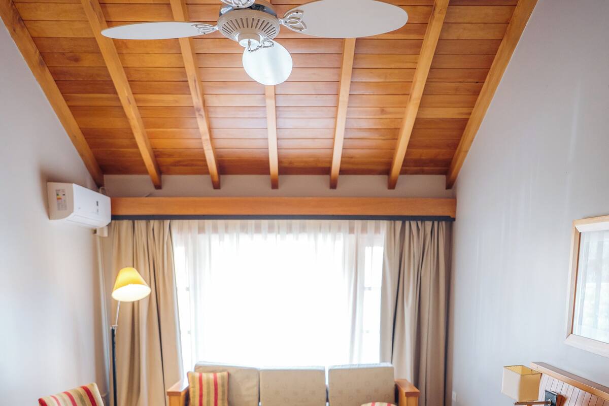 A cozy room with a wooden ceiling, a ceiling fan, a sofa, a striped armchair, a small table, and a bed with floral pillows.