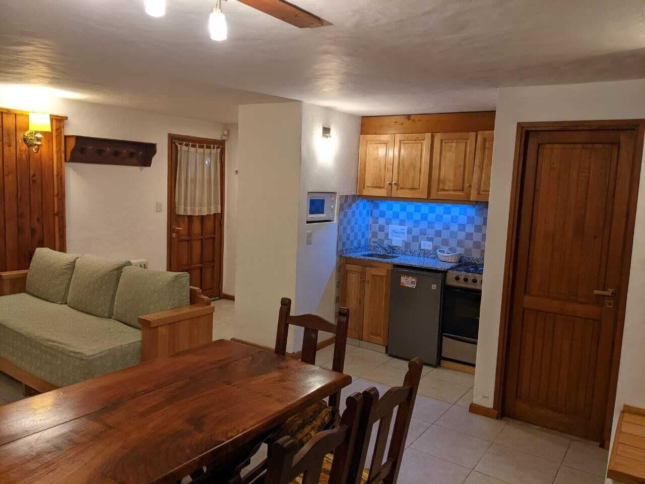 A cozy living room with a wooden dining table and chairs, a sofa, and a kitchen area with wooden cabinets and a refrigerator.