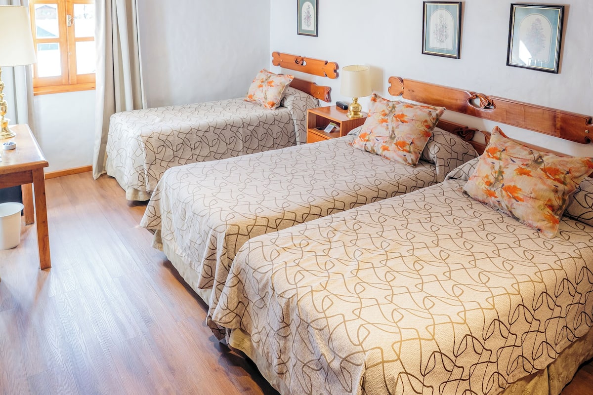 A bedroom with two beds, wooden beams, a window with curtains, and framed pictures on the wall.