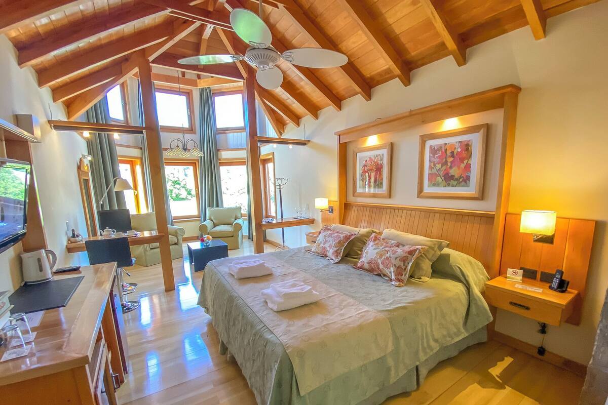 A spacious bedroom with a large bed, wooden ceiling beams, a desk with a chair, and a television.