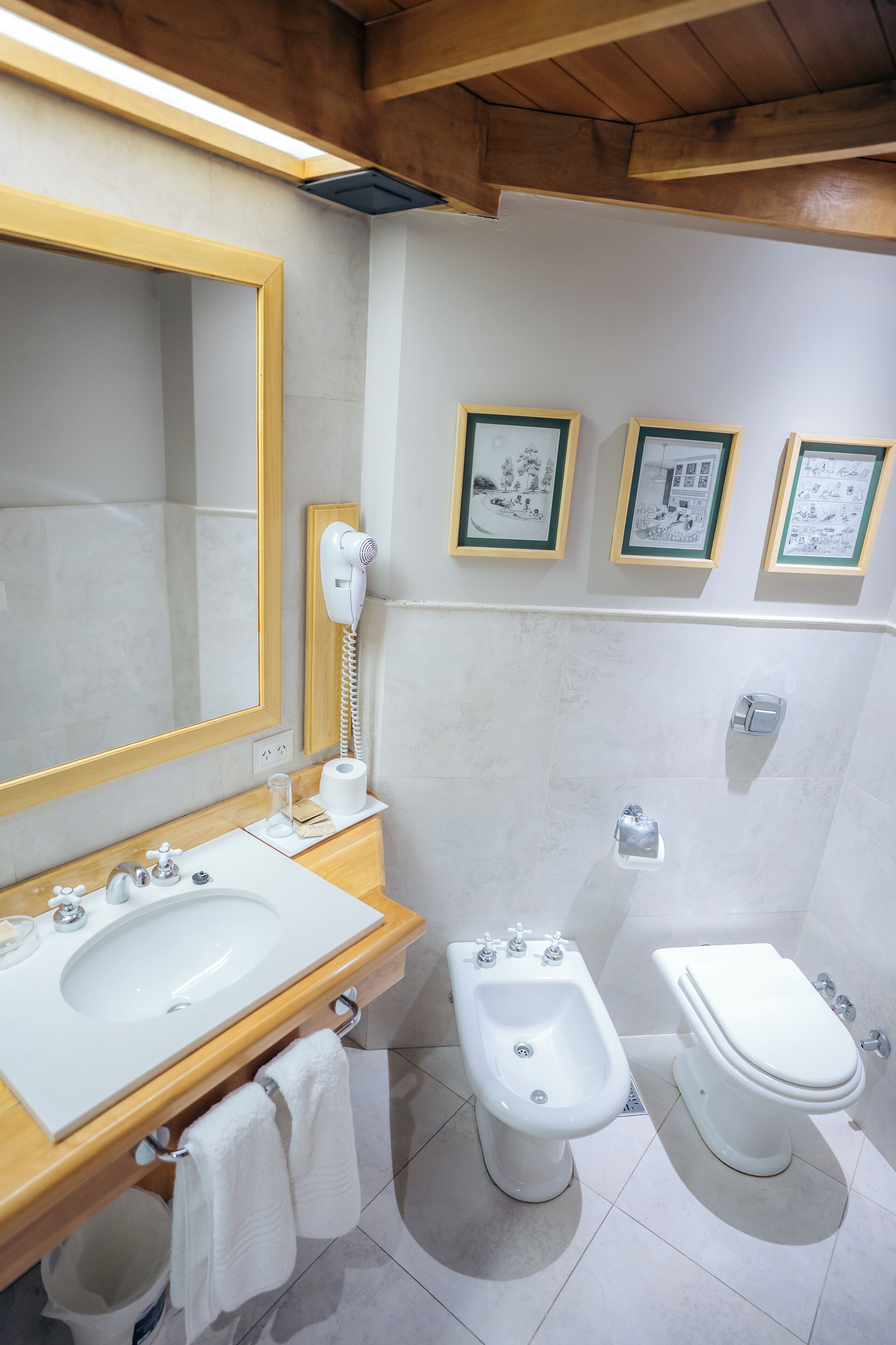Une salle de bain avec un meuble-lavabo en bois, un grand miroir, un bidet, des toilettes et des tableaux encadrés accrochés au mur.