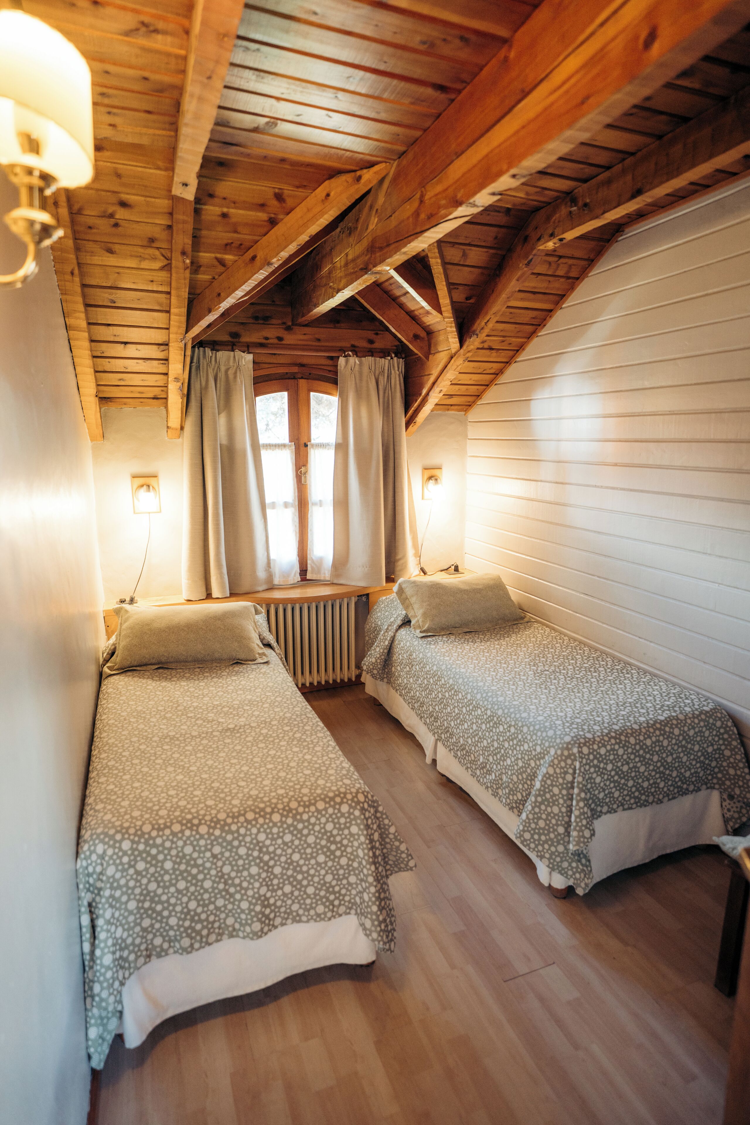 A cozy cabin room with two beds, wooden beams, a window with curtains, and a wall-mounted lamp.
