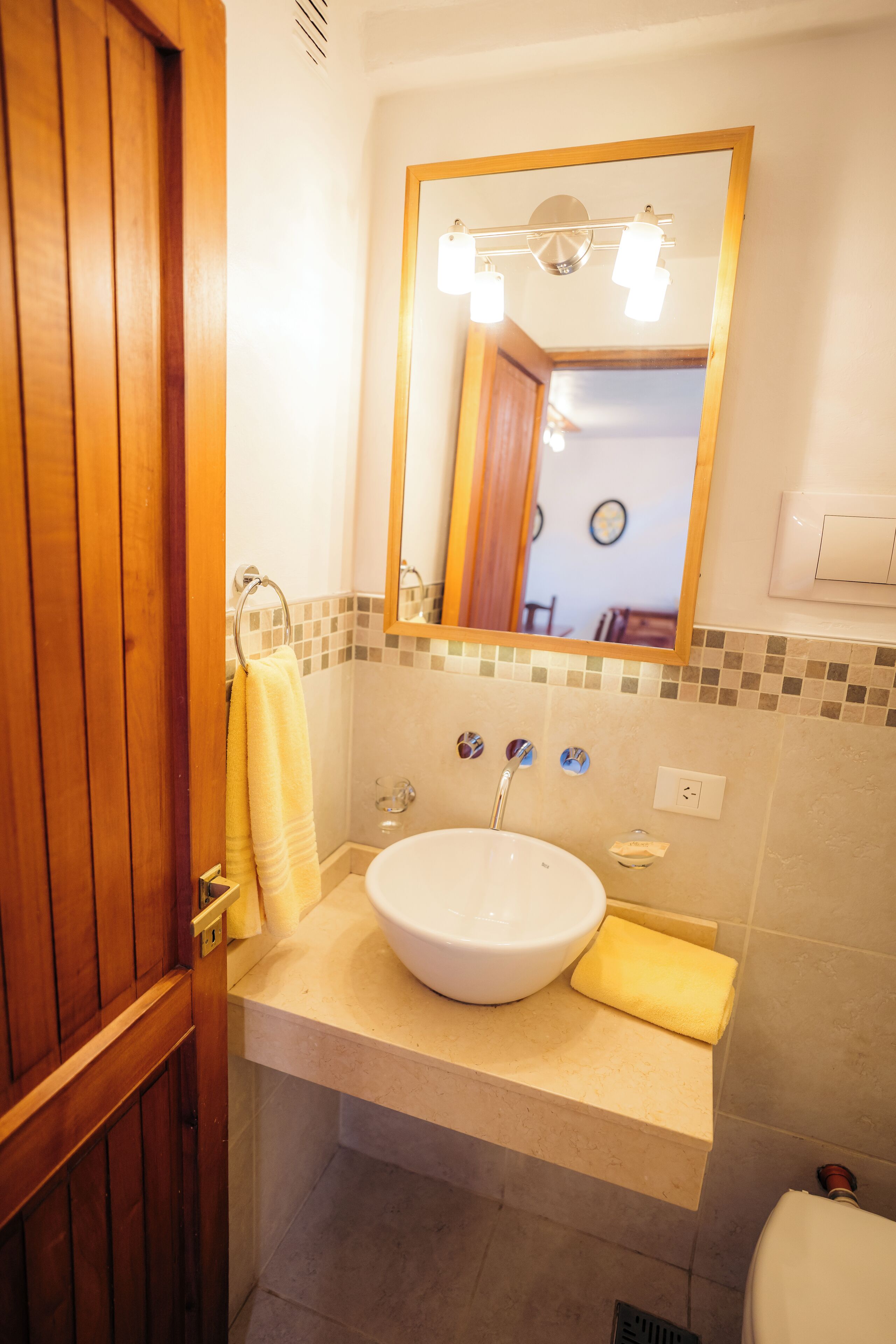 Une salle de bain avec une porte en bois, un lavabo surmonté d’un miroir et une serviette jaune accrochée au mur.