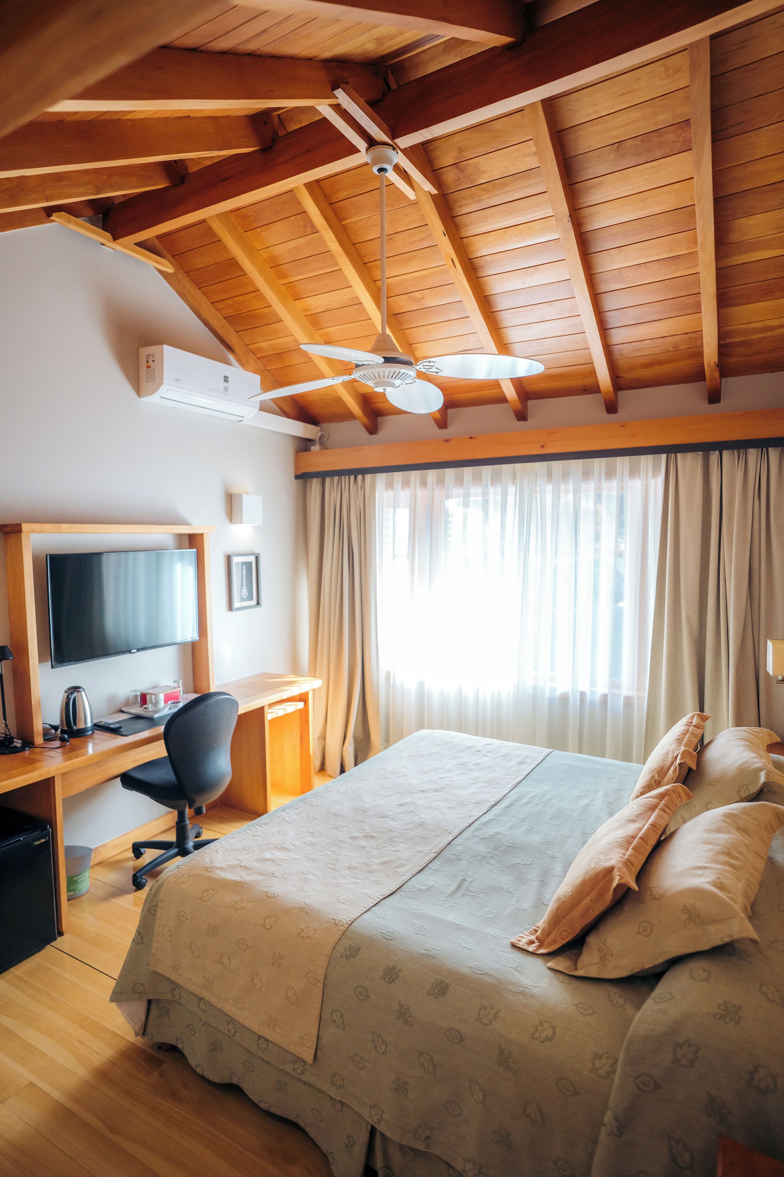 A hotel room with a wooden ceiling, a bed with a beige bedspread, a desk with a chair, a television, and a window with curtains.
