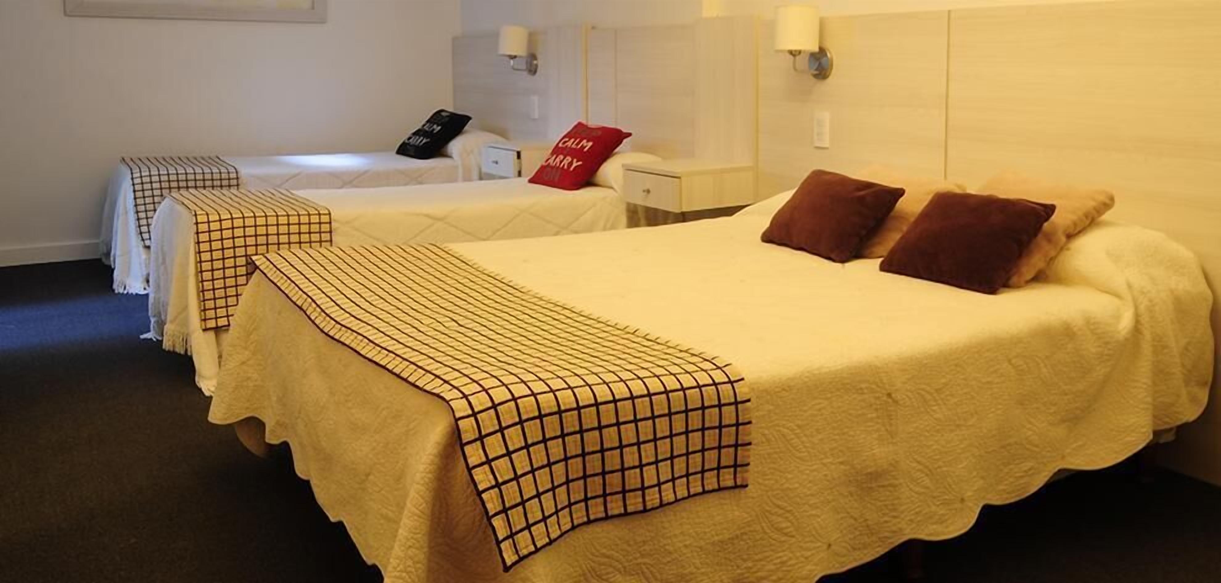 Habitación de hotel con dos camas individuales, cada una con colcha y almohadas a cuadros. Se observa un mesita de noche con una lámpara y una almohada roja con la inscripción 'CALM BARRY'.