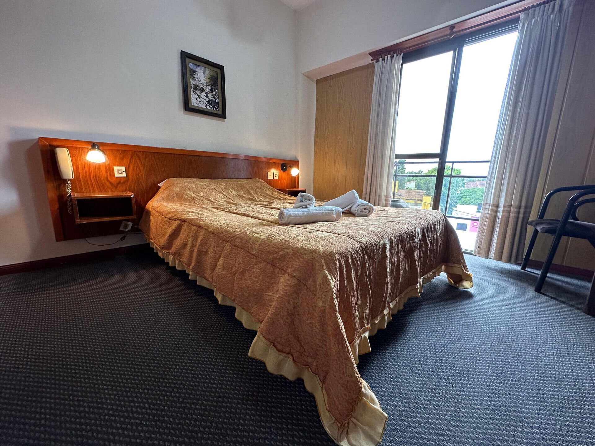 A hotel room with a large bed, a wooden headboard, a bedside telephone, and a window with curtains.