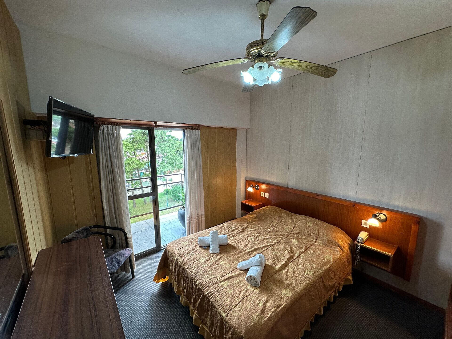 Une chambre d’hôtel équipée d’un lit, d’un bureau, d’une télévision, d’un ventilateur de plafond et d’un balcon avec des rideaux.