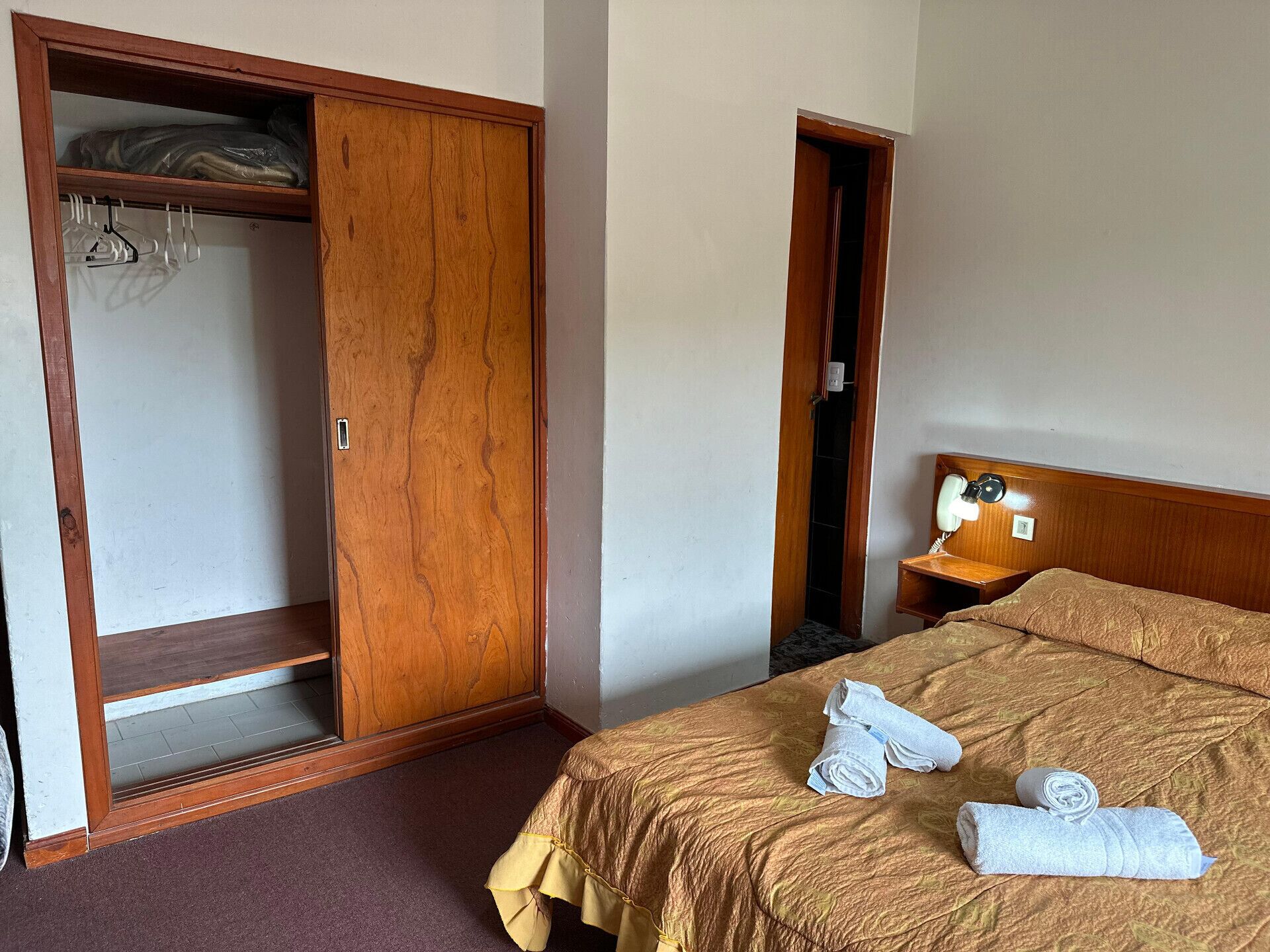 A hotel room with a wooden wardrobe, a bed with a brown bedspread, and a bedside table with a lamp and a mirror.
