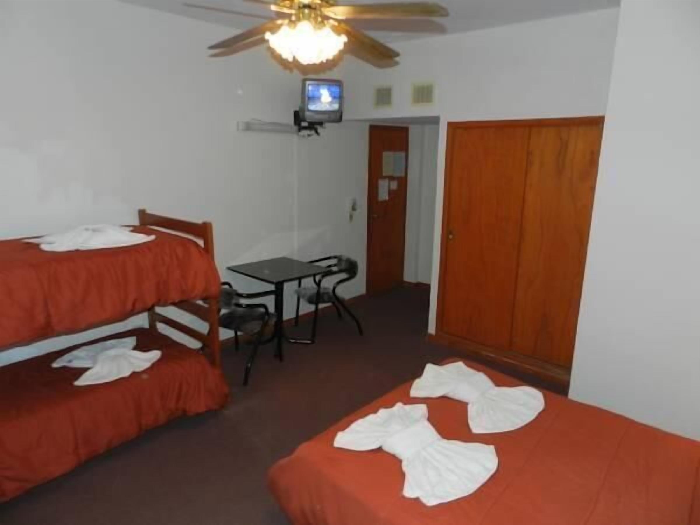 A room with two single beds, a small table, a chair, a ceiling fan, and a television on a shelf.