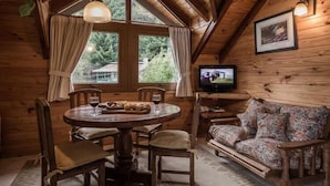 A cozy cabin interior with a wooden dining table, chairs, a sofa, a television, and a large window with curtains. - Cabañas y Apart Antuen (San Martin de los Andes)