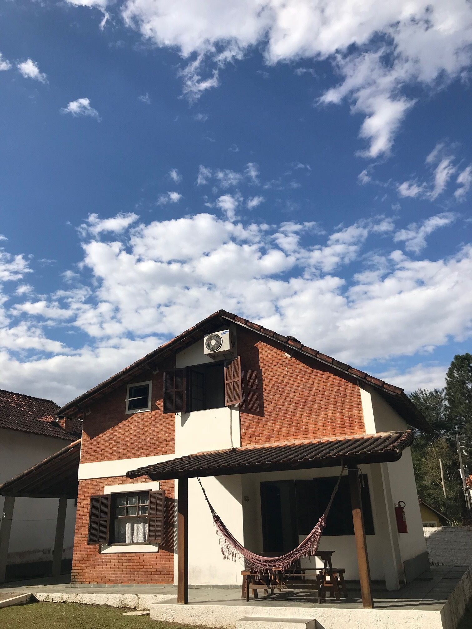 A two-story house with a brick facade, a balcony with a hammock, and a clear blue sky with clouds.