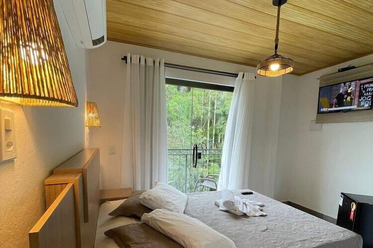 Une chambre à coucher comprenant un lit, des tables de chevet, un luminaire suspendu, une télévision et une fenêtre avec des rideaux.