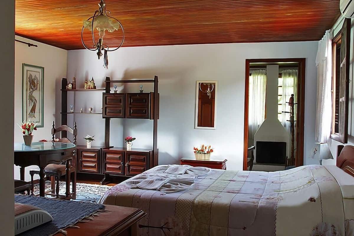 Une chambre avec un plafond en bois, un lit recouvert d’une couvre-lit à motifs floraux, une commode en bois, une chaise, une table ornée d’un vase de fleurs et une cheminée.