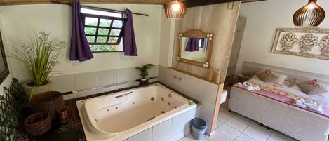 A bathroom with a bathtub, a shower, a sink, and a mirror. There is a window with purple curtains, a potted plant, and a trash can.
