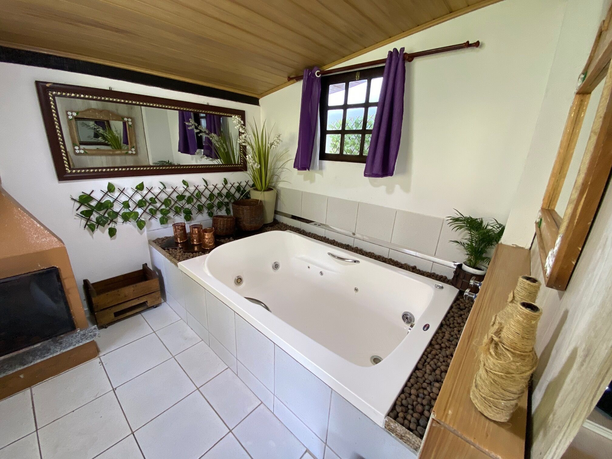 A bathroom with a large bathtub, a mirror, a window with purple curtains, and a potted plant.