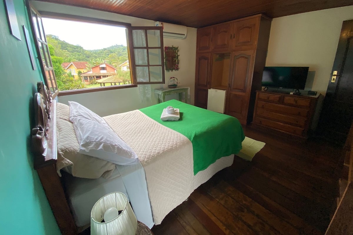 Une chambre à coucher avec un lit, une couverture verte, un téléviseur, des meubles en bois et une fenêtre donnant sur des maisons.