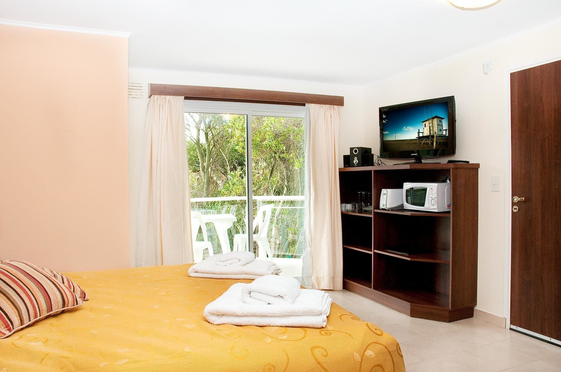 A bedroom with a bed, a TV on a wooden shelf, and a view of greenery outside.