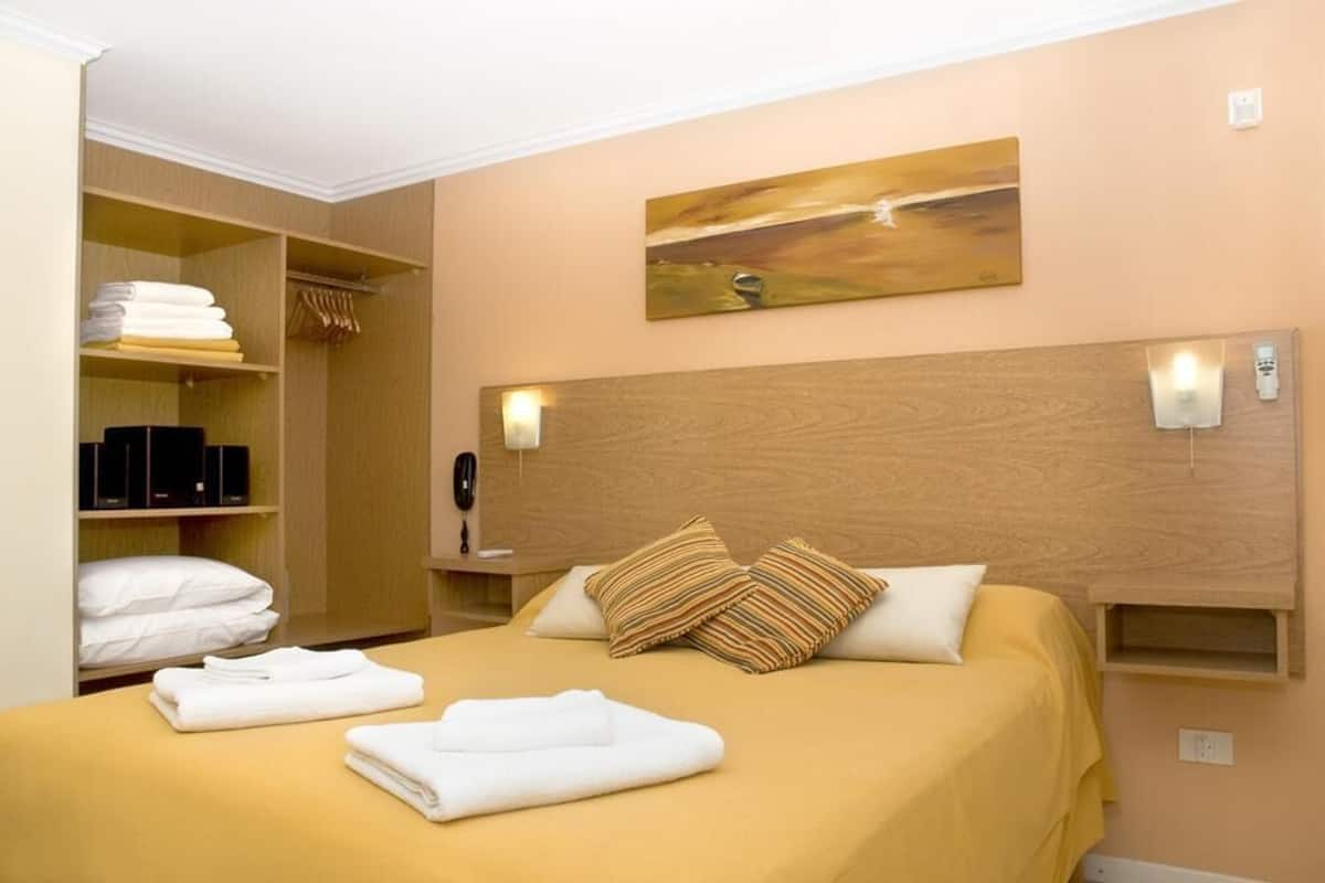 A hotel room with a bed, a wooden headboard, a painting on the wall, and a closet with folded towels.