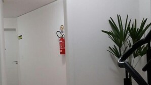A hallway with a wooden floor, white walls, and a black railing. There is a fire extinguisher mounted on the wall and a potted plant on the right side.