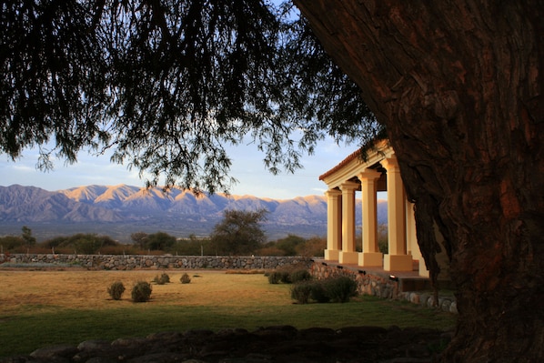 Un edificio clásico con columnas, rodeado por un muro de piedra y un área verde con un árbol.