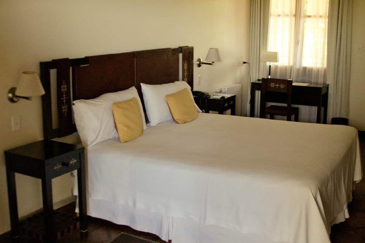 Una cama bien tendida con ropa de cama blanca y beige, un cabecero de madera y dos mesitas de noche con lámparas.