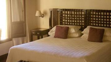 Una cama bien hecha con cabecera de madera, una mesita de noche con lámpara y una ventana con cortinas.
