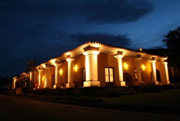 A two-story building with a symmetrical facade, featuring a central entrance flanked by columns, and illuminated by warm exterior lighting.