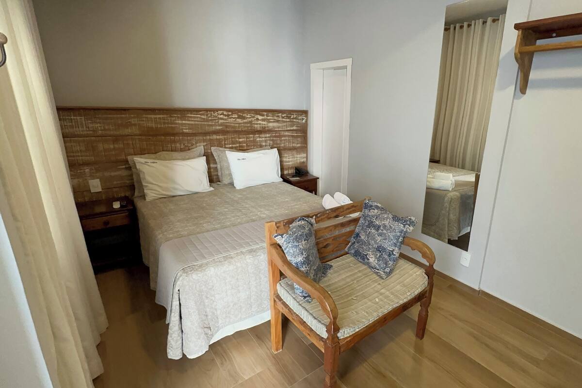 A bedroom with a wooden headboard, a bed with white linens, a wooden chair with a cushion, and a mirror on the wall.