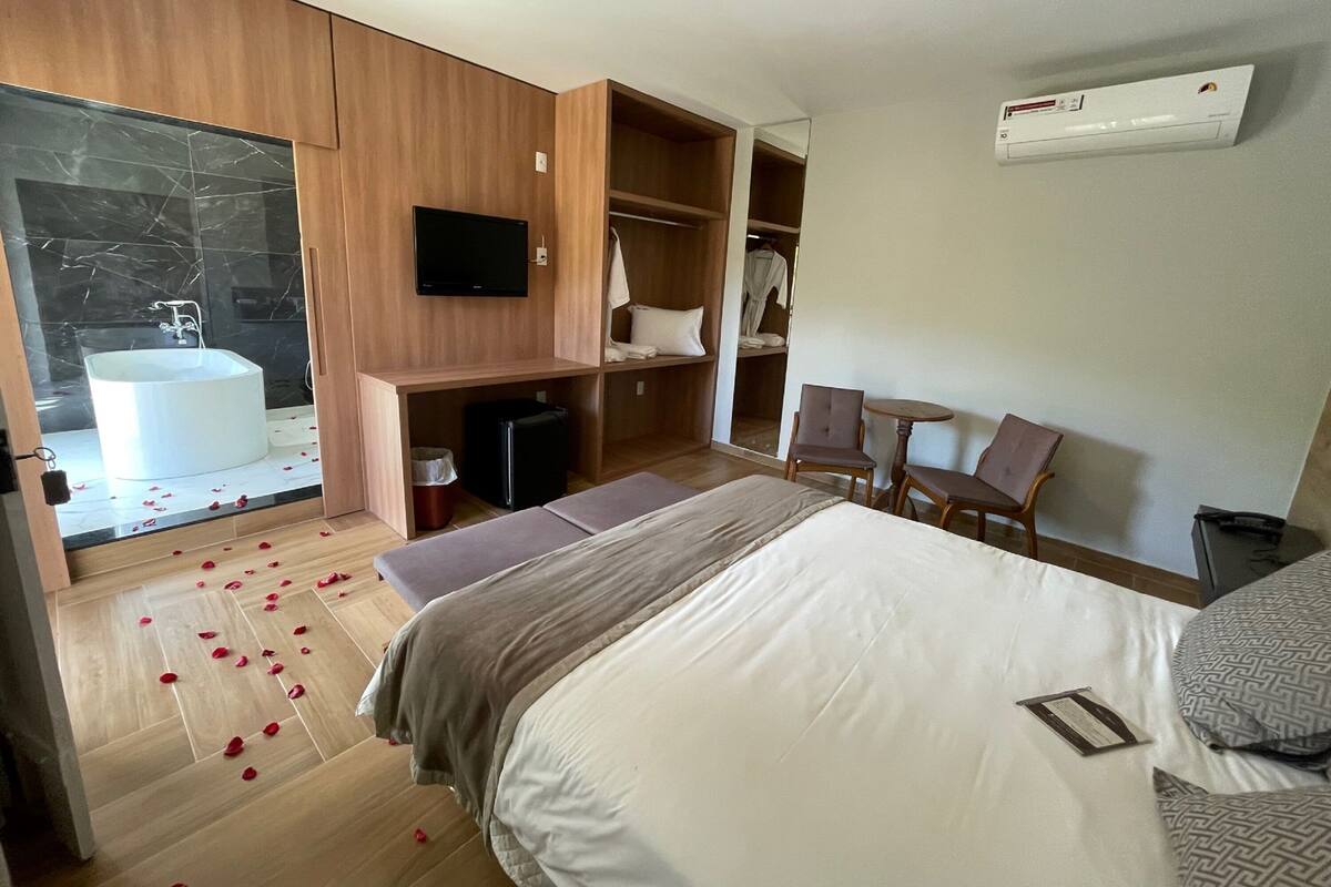 A modern hotel room with a large bed, a built-in TV, a bathroom with a bathtub, and a seating area with chairs and a table.