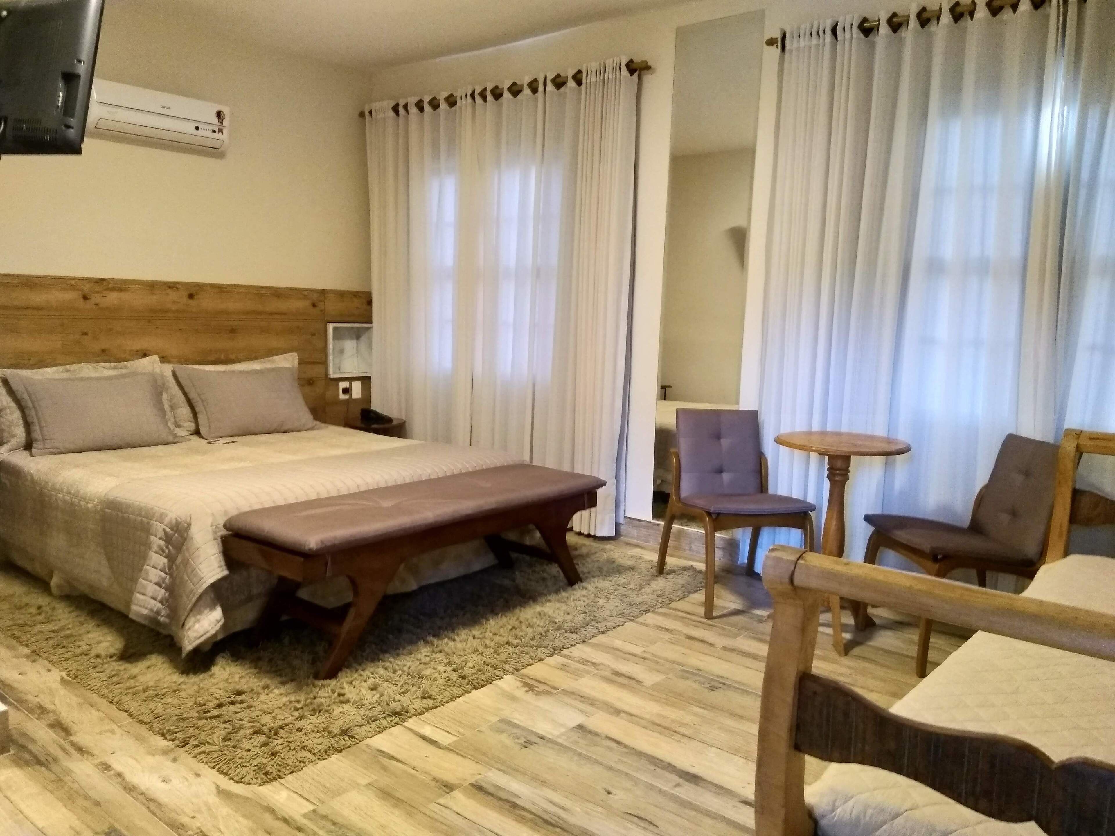 A hotel room with a wooden headboard, a large bed with a bench, two chairs, a small round table, and a TV mounted on the wall.