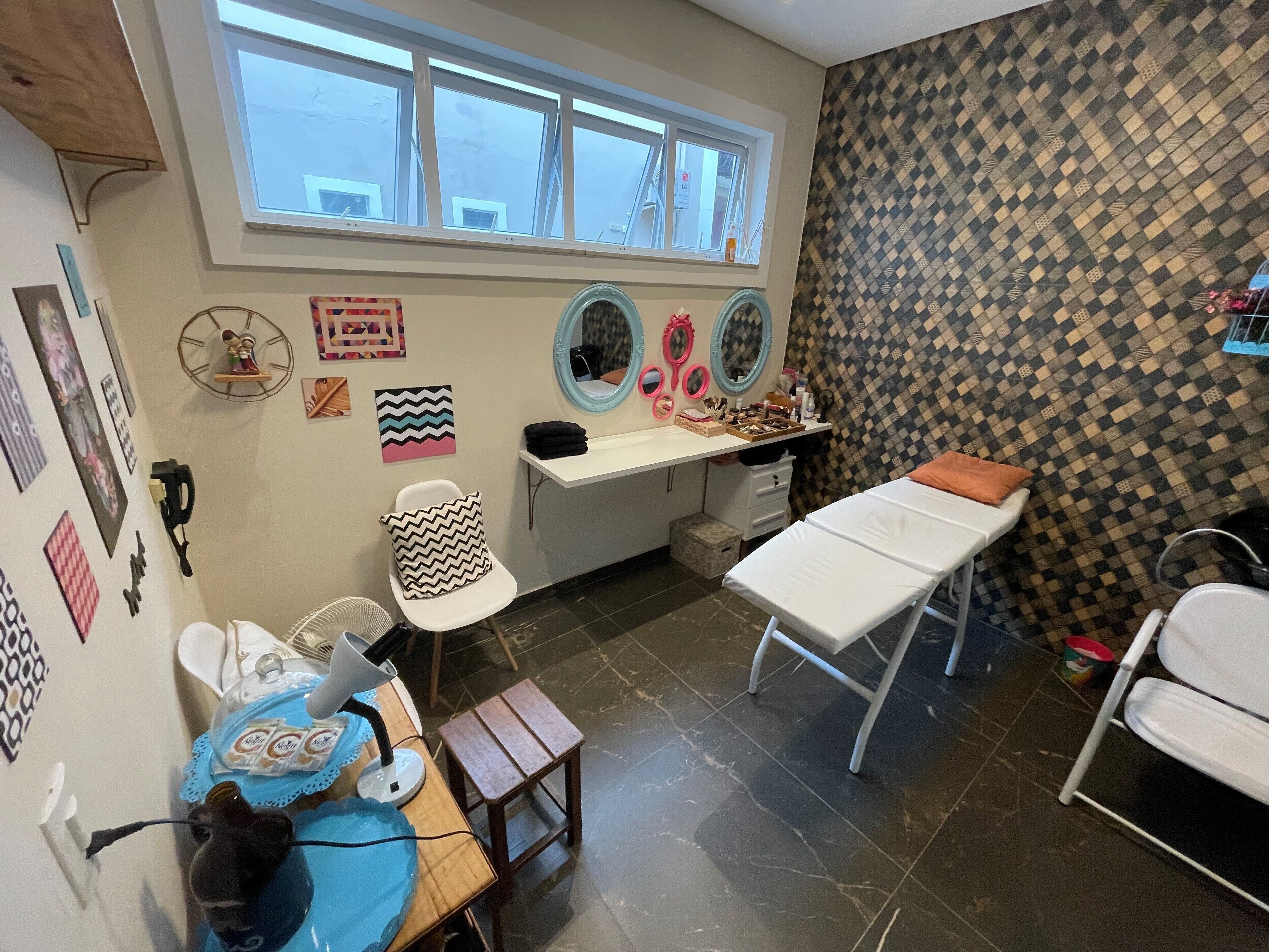 A room with a large window, a vanity with mirrors, a small table with a lamp, and a bed.