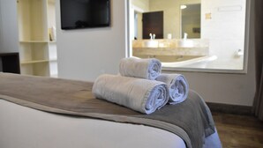 Una habitación de hotel con una cama, un televisor en la pared y un baño visible a través de un ventanal grande.