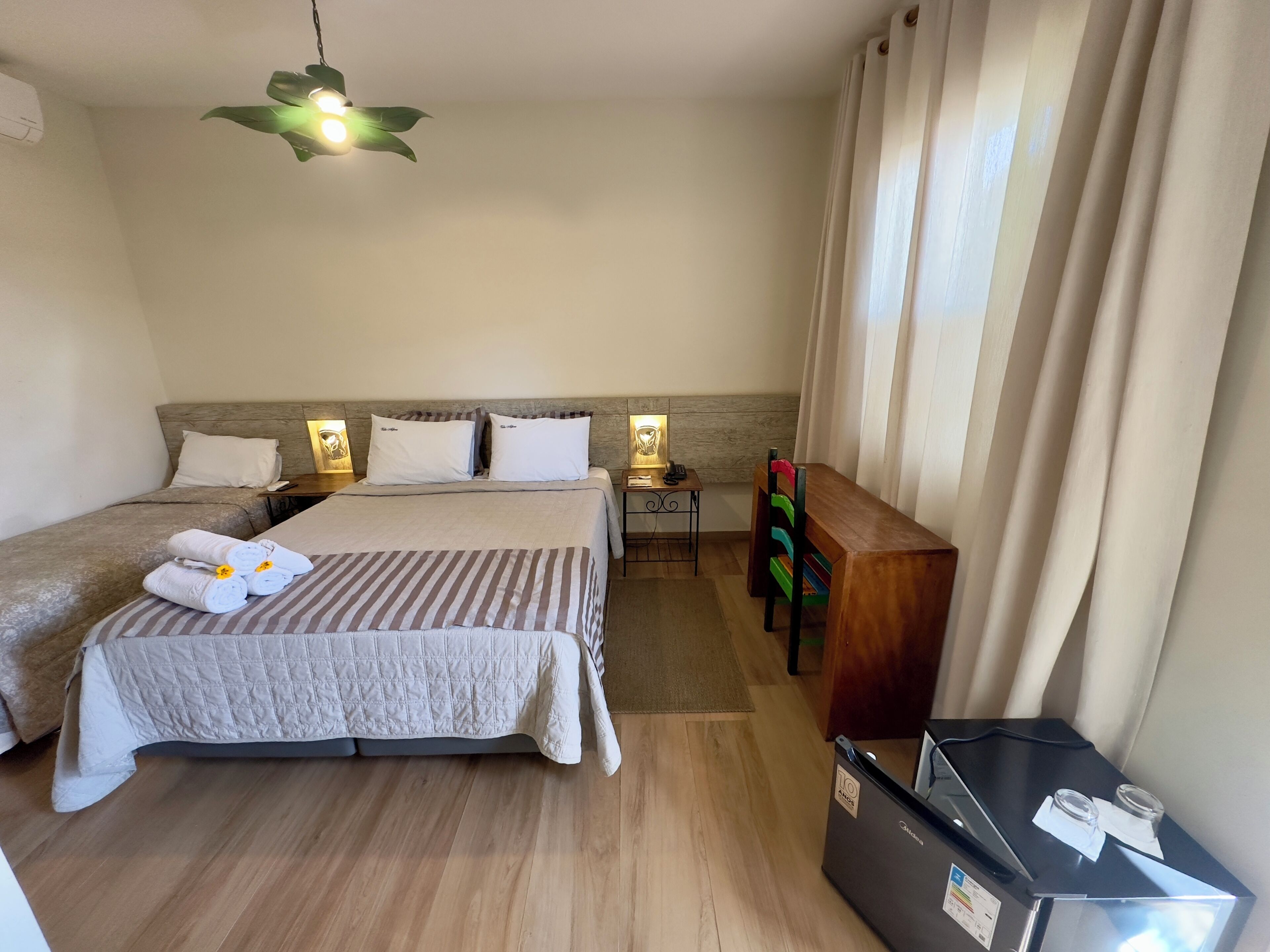 A hotel room with a bed, bedside tables, a desk, a chair, a desk lamp, a refrigerator, and curtains.