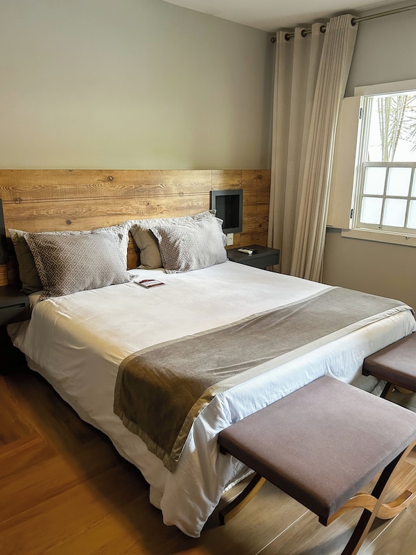 Una cama bien hecha con cabecera de madera, una mesita de noche con un control remoto y una ventana con cortinas.