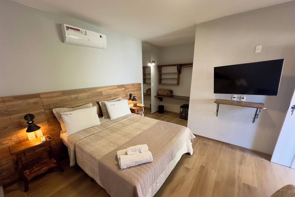 A hotel room with a wooden headboard, a bed with a beige quilt, a bedside lamp, a flat-screen TV mounted on the wall, and a wooden floor.