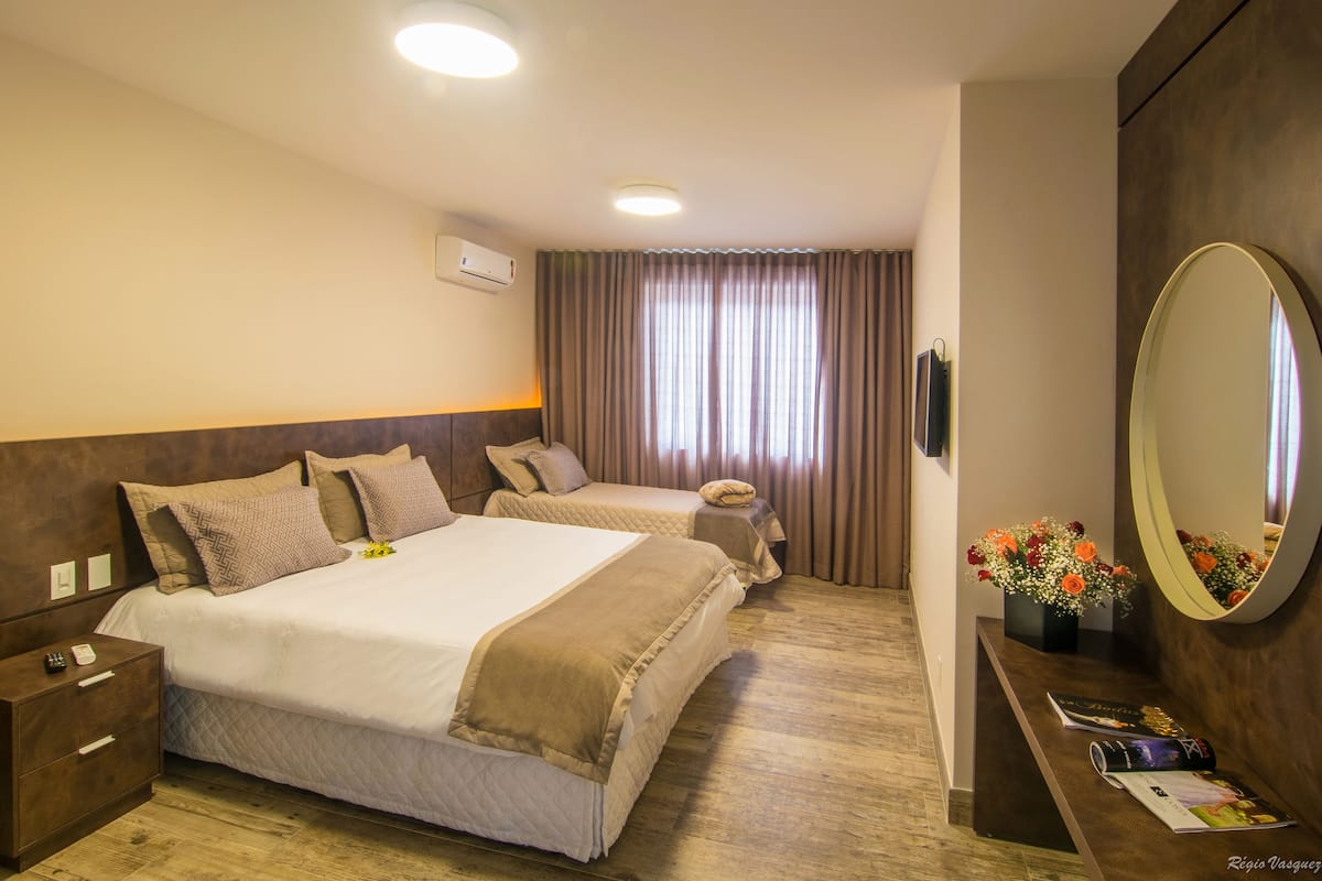 A hotel room with two beds, a bedside table with a lamp, a mirror, and a window with curtains.