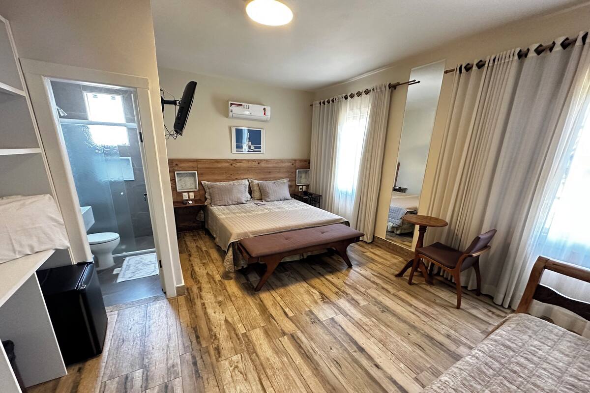 A hotel room with a wooden floor, a bed with a bench, a bedside table, a chair, a small table, a mirror, an air conditioning unit, and a window with curtains.
