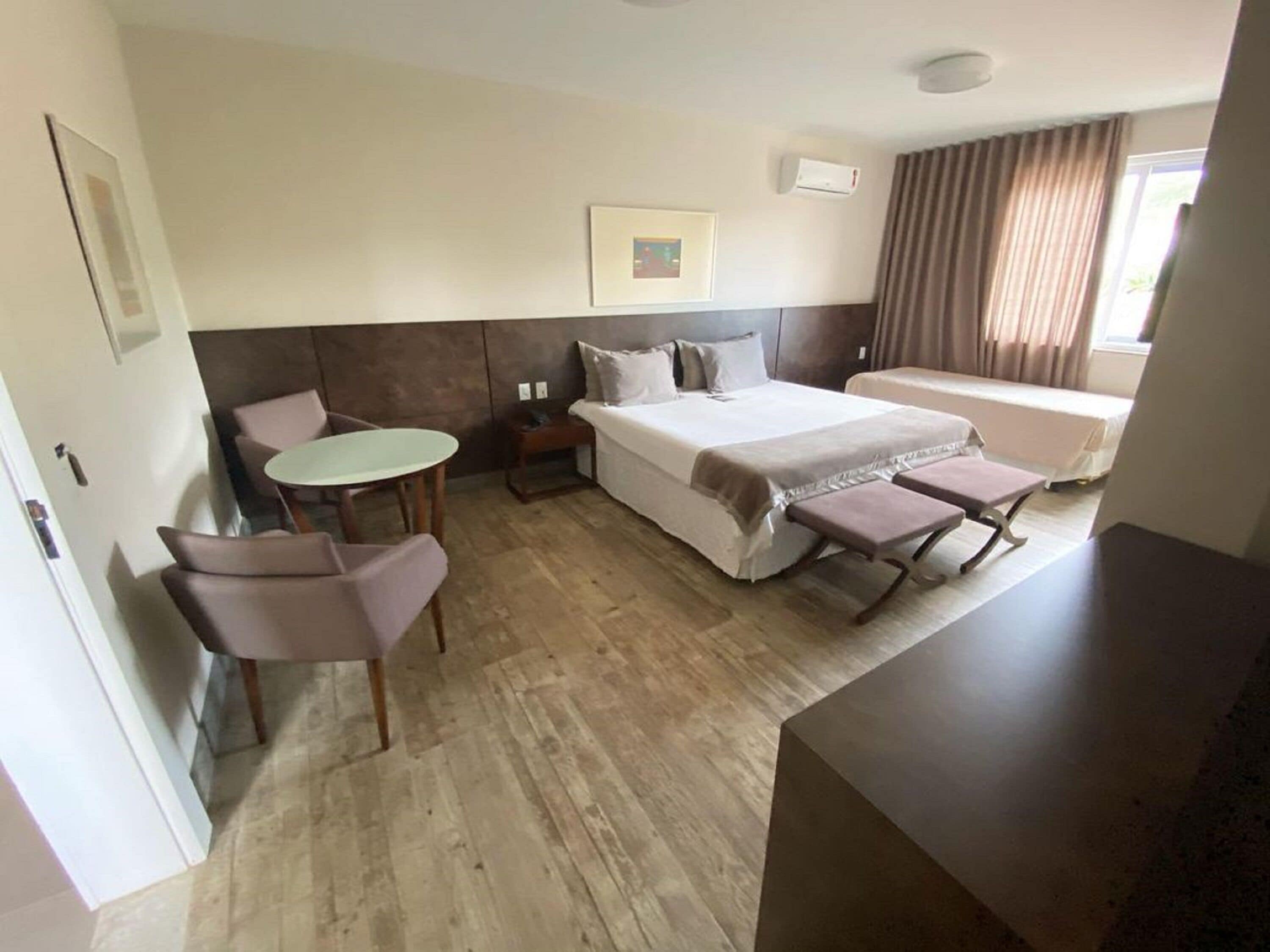 A hotel room with a bed, a small table with two chairs, a bedside table, and a window with curtains.