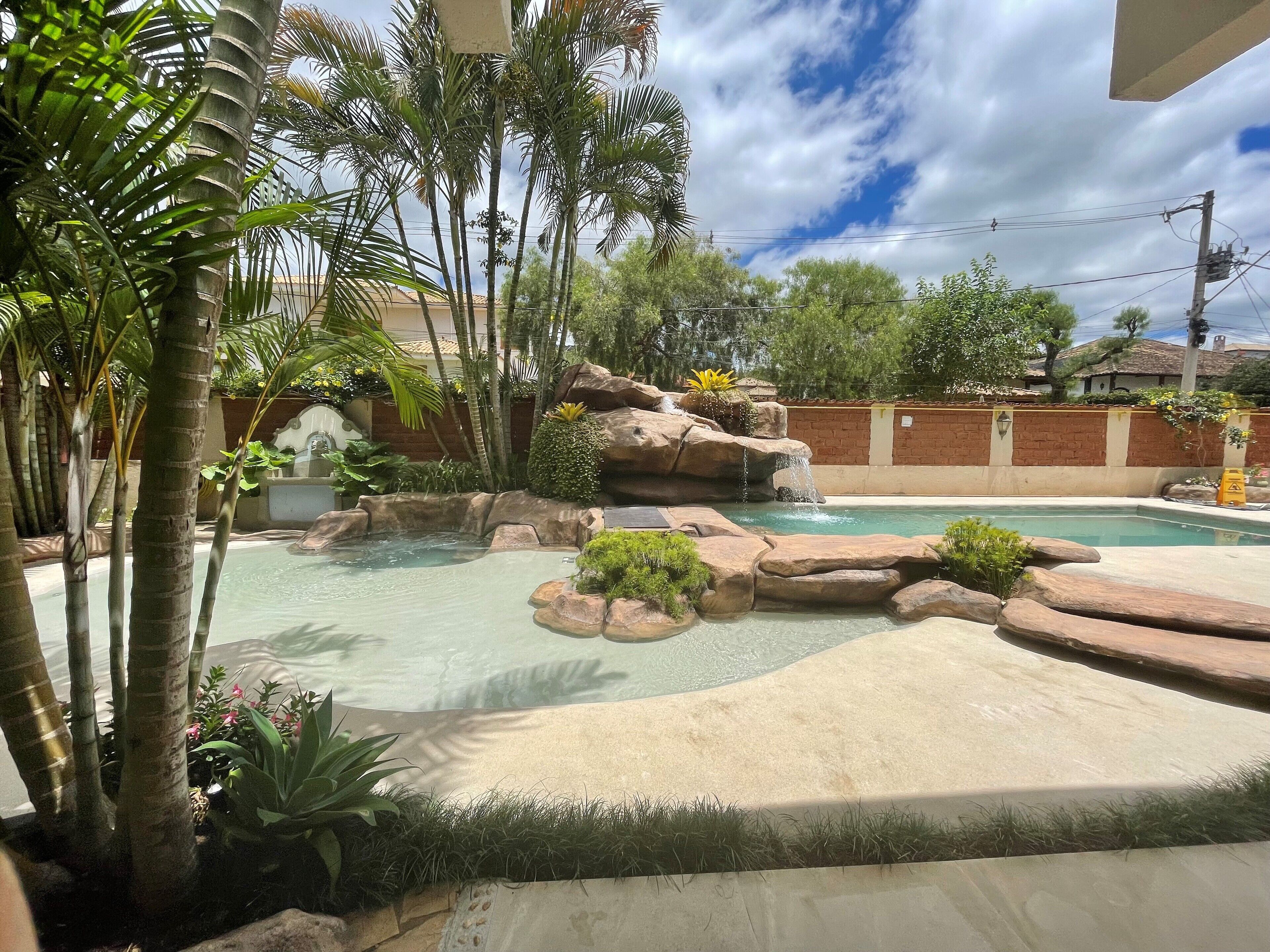 Une piscine au design naturel, avec des rochers, entourée de plantes tropicales et sous un ciel dégagé.