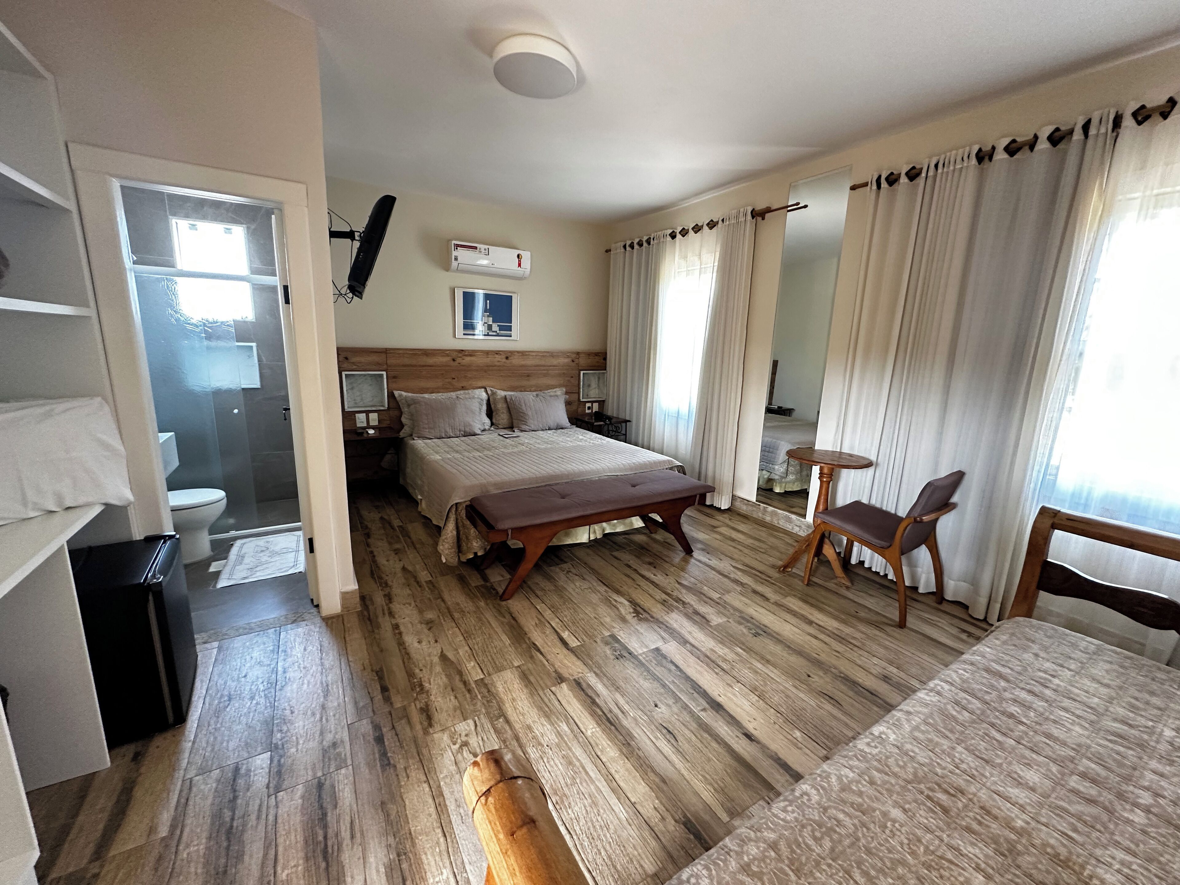 A hotel room with a wooden floor, a large bed with a headboard, a bedside table, a chair, and a small table. There is a window with curtains, a door leading to the bathroom, and an air conditioning unit on the wall.