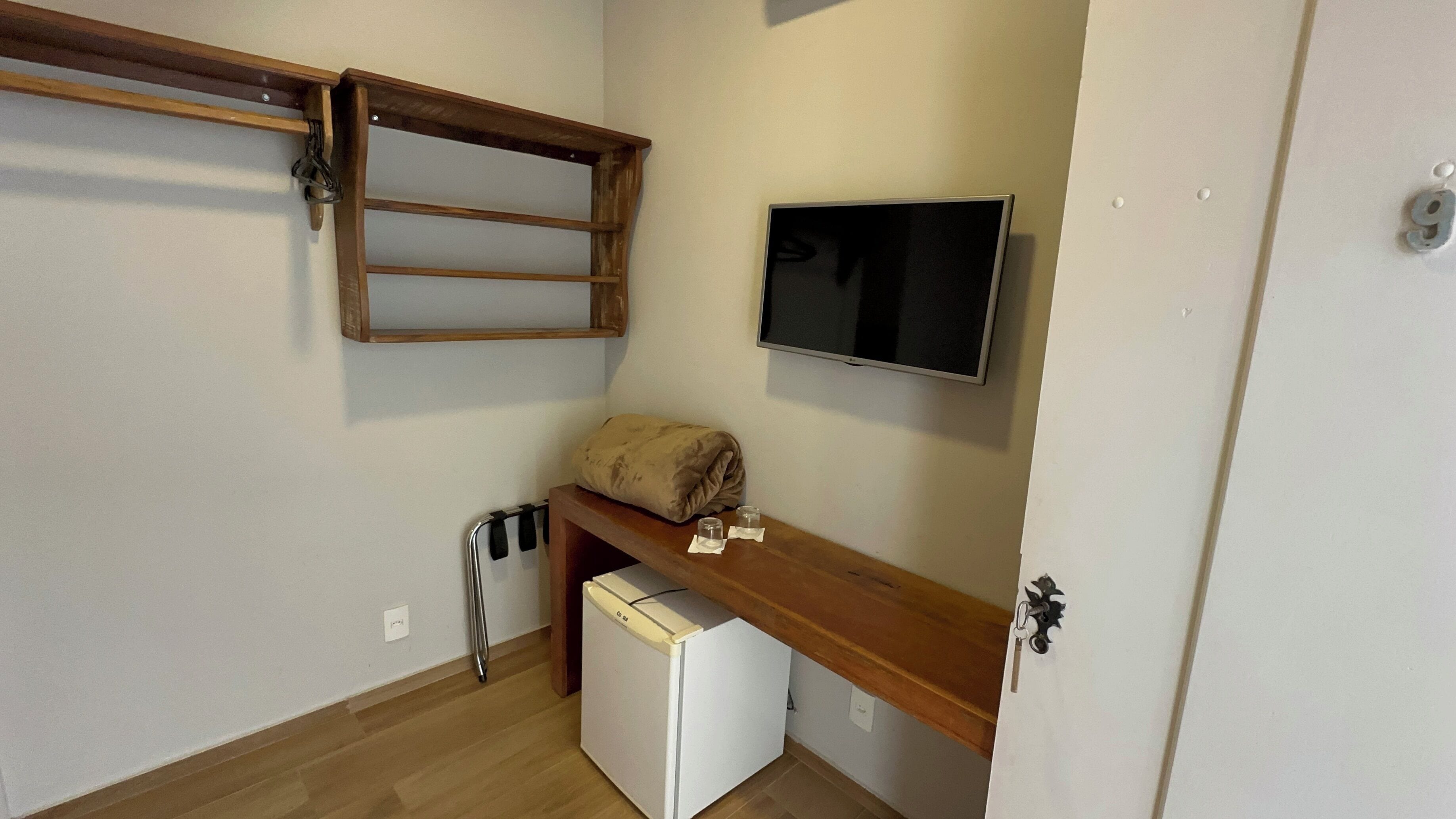 Habitación pequeña con un banco de madera, un refrigerador blanco y una televisión montada en la pared, y una puerta con cerradura.