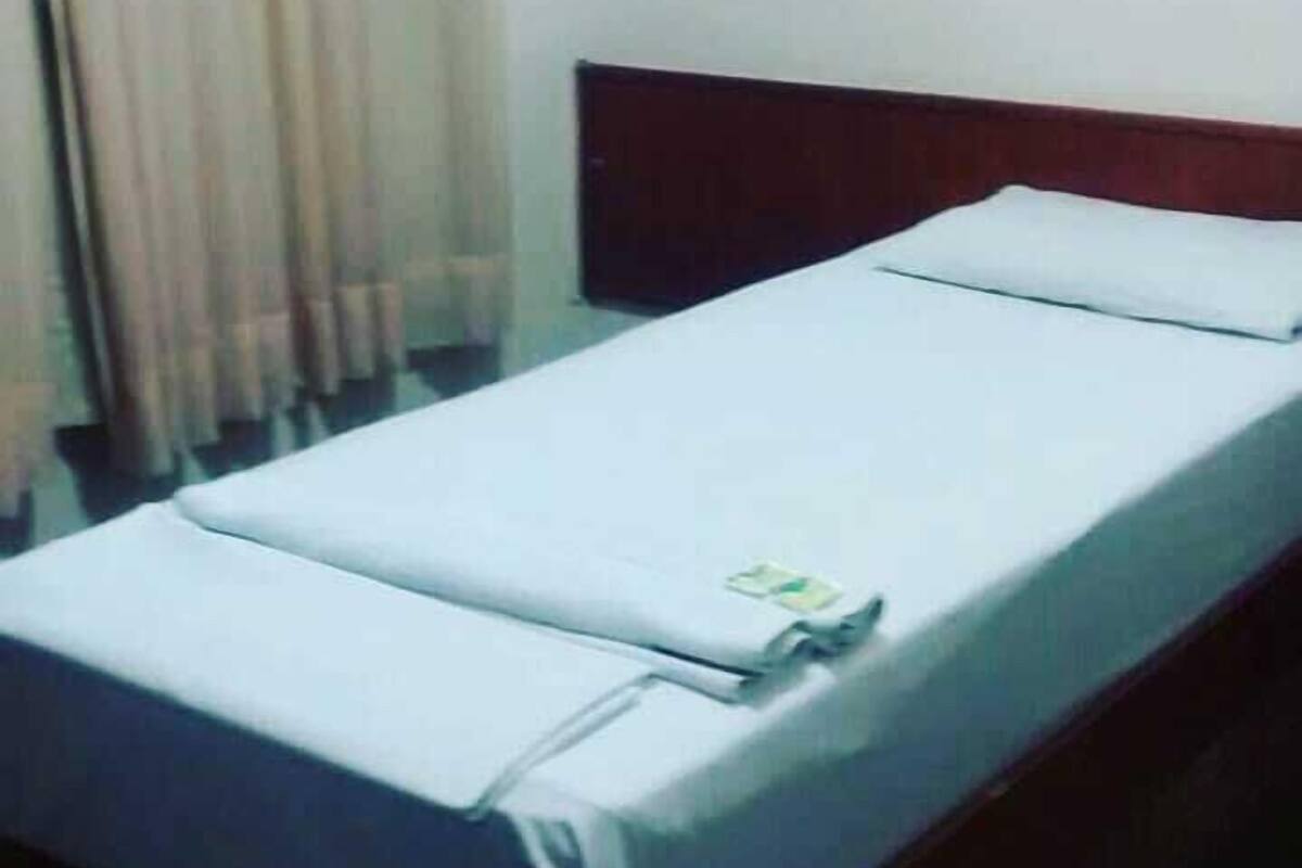 A single bed with a wooden headboard in a room with a plain white wall and a window with curtains.