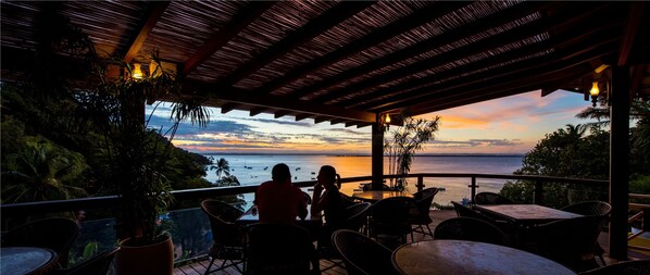 An outdoor seating area with tables and chairs, overlooking a body of water and a sunset.