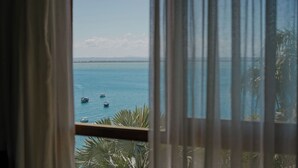 Blick durch ein Fenster mit Vorhängen, der ein ruhiges Meer mit mehreren Booten und einer fernen Küste zeigt.