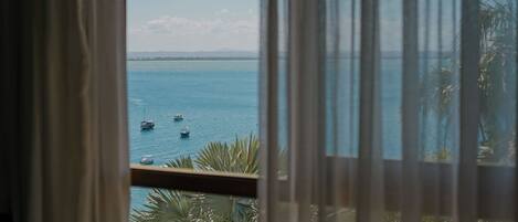 Vista a través de una ventana con cortinas, mostrando un mar tranquilo con varios barcos y una costa lejana.