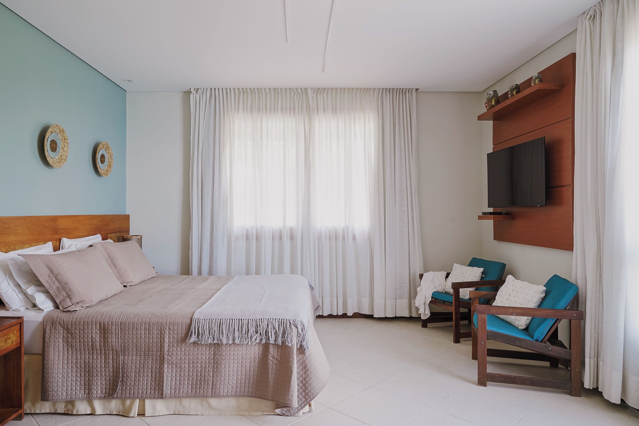 Une chambre à coucher avec un lit, deux chaises, une télévision et une fenêtre avec des rideaux.
