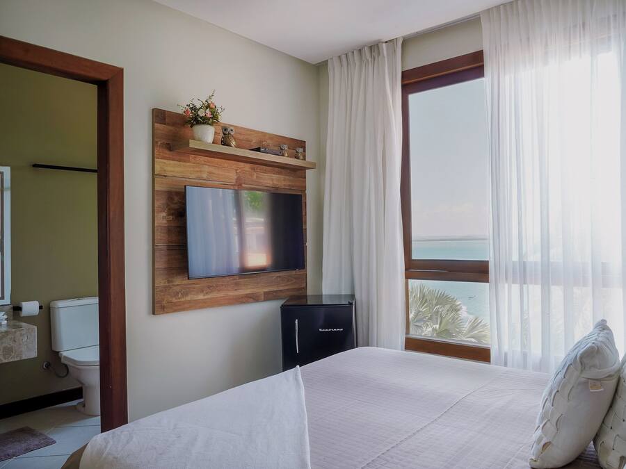 A hotel room with a bed, a TV mounted on a wooden panel, a small black refrigerator, a window with a view of the ocean, and a bathroom visible through an open door.