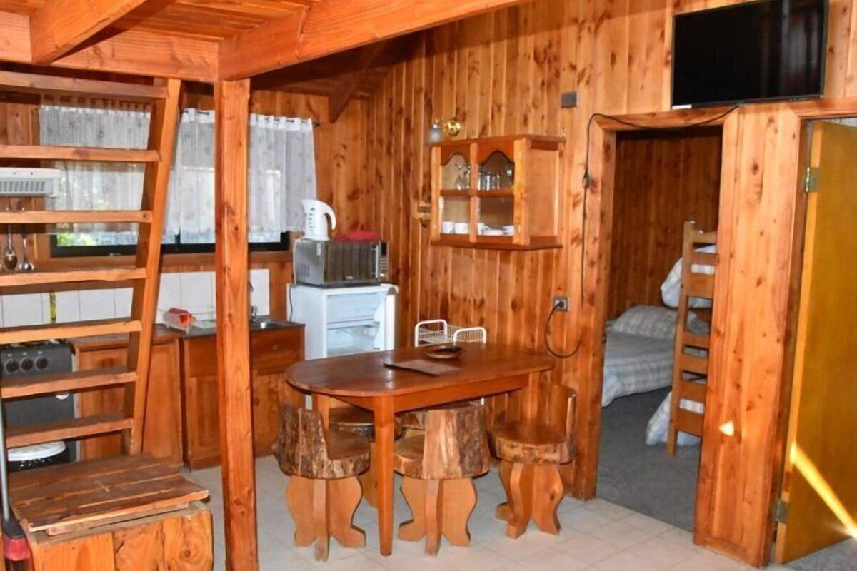 A wooden interior with a dining table, chairs, a kitchen area with appliances, and a television mounted on the wall.