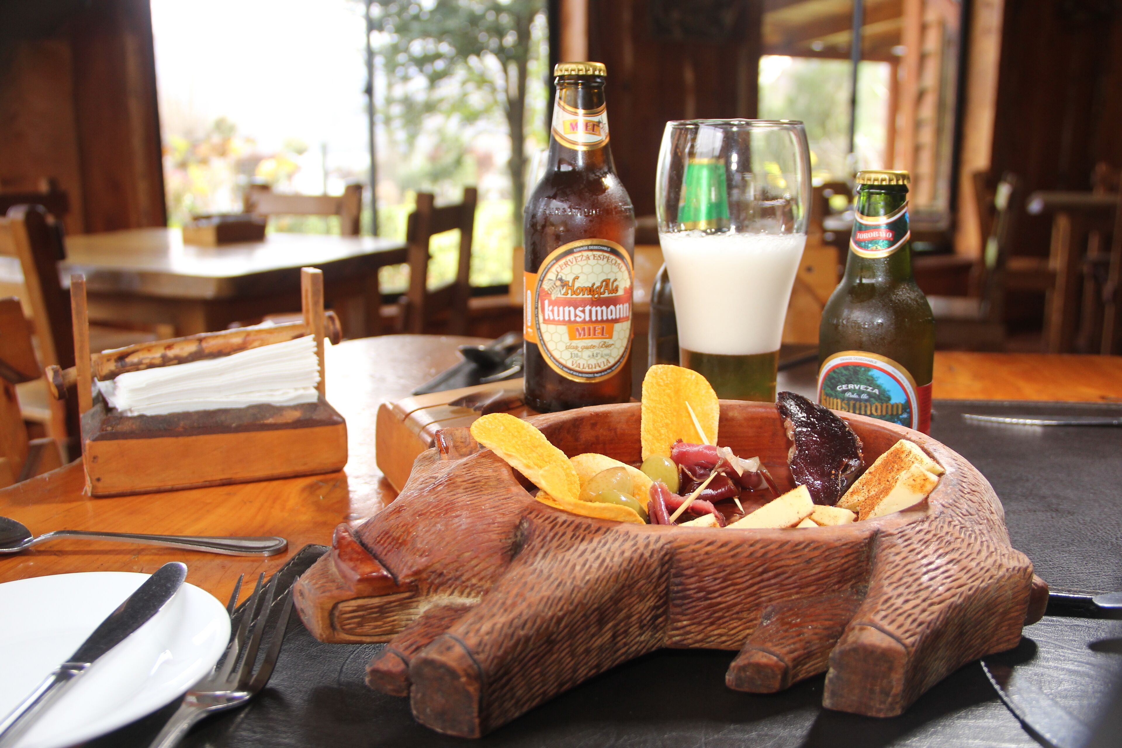 Un cuenco con forma de cerdo de madera lleno de aperitivos y dos botellas de cerveza sobre una mesa.